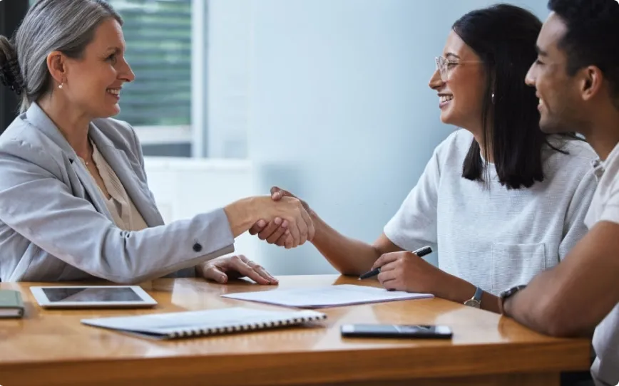 A couple shakes hand with a woman over a table of papers.