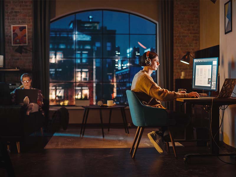 Two people are working on computers while staying in an apartment loft in the city. One person sits on the couch while another has a desk with a large monitor. They could be airbnb guests or tenants of the home as it's not easy to tell.